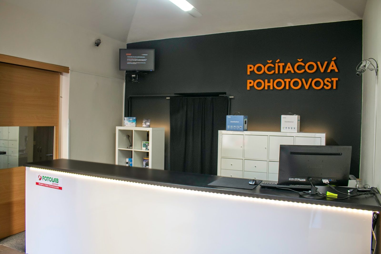 Příjmový pult provozovny s prosvětlenou čelní deskou, za ním černá stěna s oranžovým logem POČÍTAČOVÁ POHOTOVOST a police s příslušenstvím